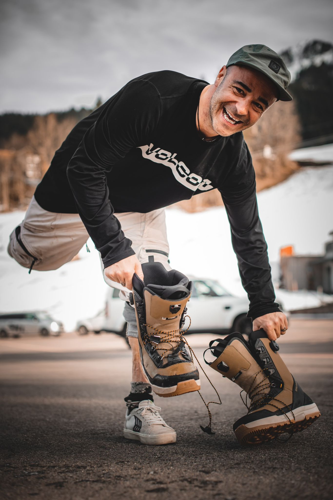 Orlando, founder of Orlando Projects, smiling and holding two snowboard boots in front of a snowy mountain backdrop. Wearing a black Volcom long-sleeve shirt and a green cap.